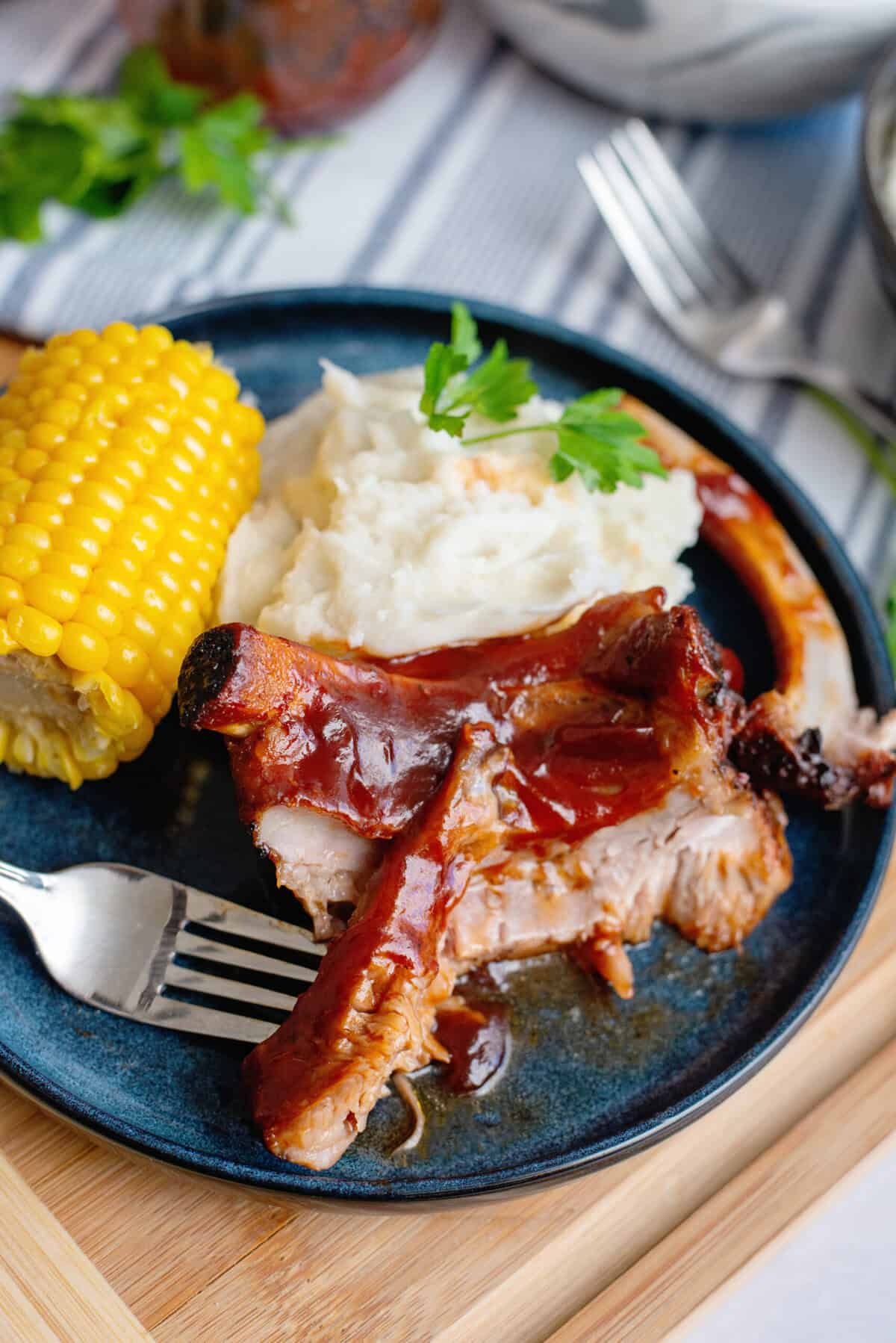 A close-up of a barbecue meal on a dark blue plate, featuring tender, saucy ribs with a deep red glaze, creamy mashed potatoes garnished with a sprig of fresh parsley, and a bright yellow ear of corn on the cob. A silver fork rests on the plate, with a piece of rib meat resting on its tines. The background includes a striped cloth napkin, a glass jar, and additional tableware. The setting is warm and inviting, emphasizing comfort food.