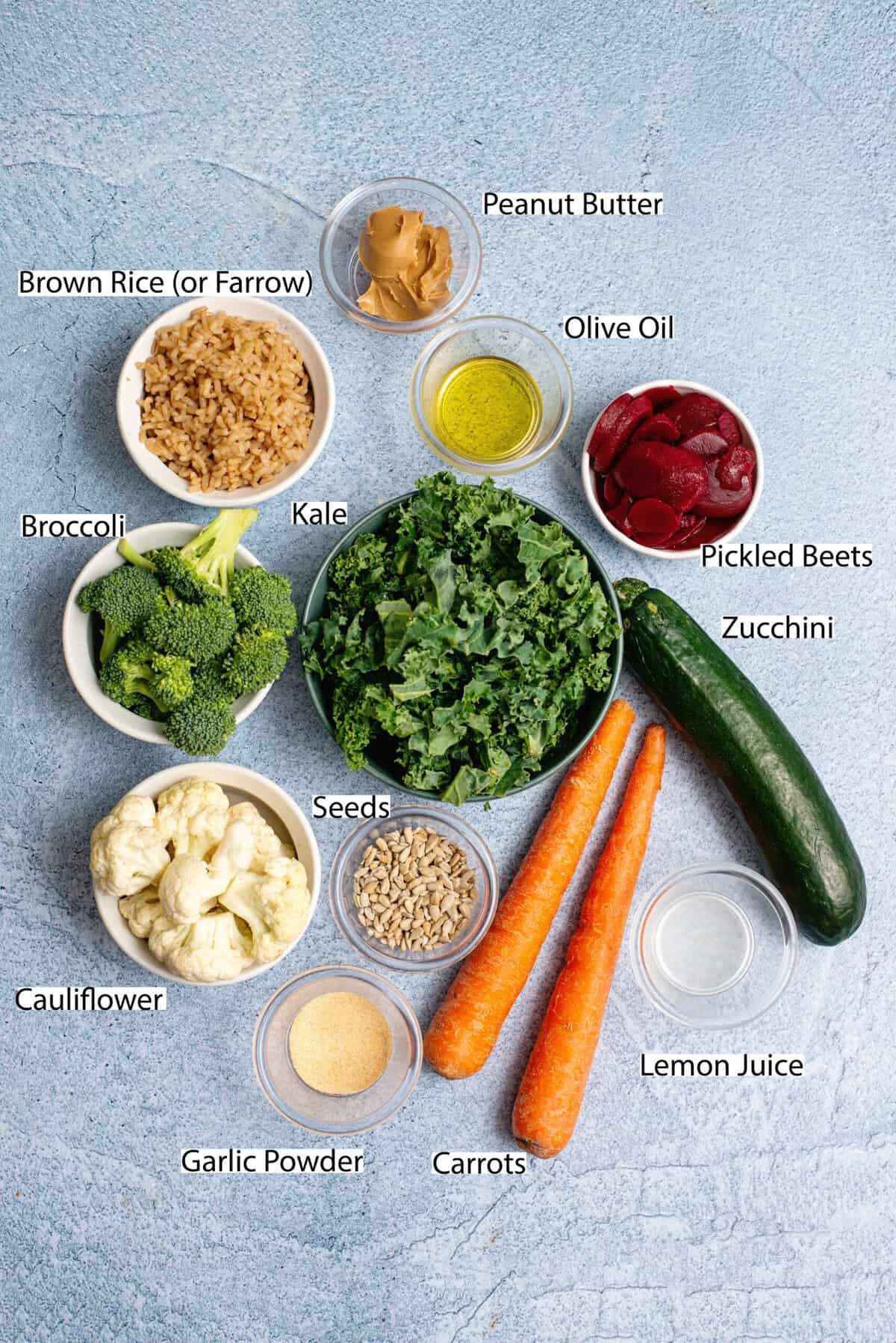 An assortment of fresh ingredients arranged on a textured blue background, including kale, carrots, zucchini, broccoli, cauliflower, brown rice, beets, peanut butter, sunflower seeds, olive oil, and seasonings, ready for a nutritious grain bowl.