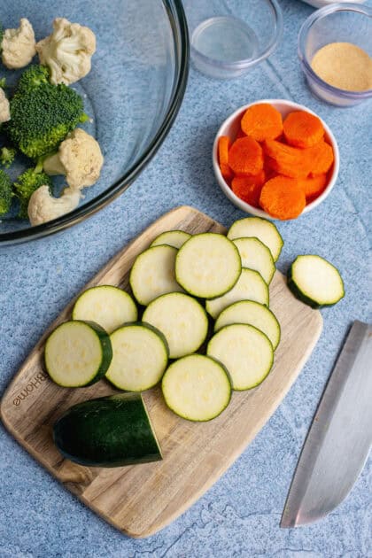 A cutting board with freshly sliced zucchini, surrounded by a bowl of chopped carrots, a mixing bowl with broccoli and cauliflower, and various seasonings on a blue countertop.