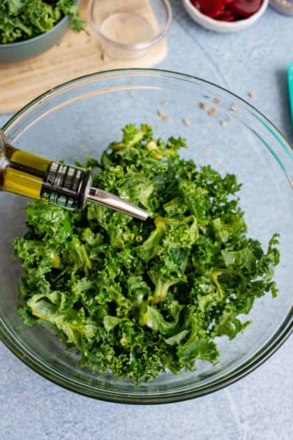 A close-up of fresh kale in a glass bowl being drizzled with olive oil, with additional ingredients like beets and sunflower seeds in the background.