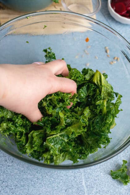 A hand gently massaging fresh kale in a glass mixing bowl, softening the leaves to make them tender and flavorful for a salad or grain bowl.