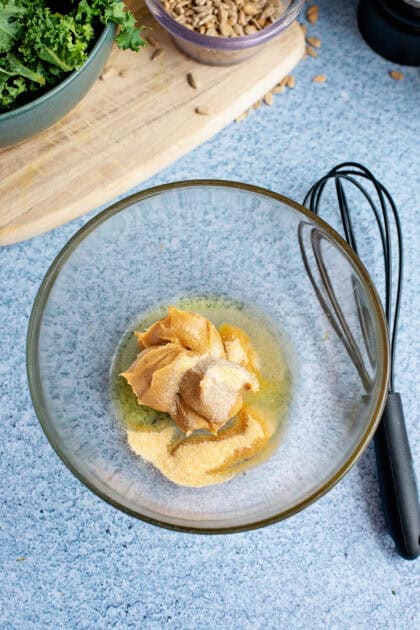 A glass bowl with peanut butter, olive oil, and seasonings, ready to be whisked into a creamy peanut dressing. A whisk and additional ingredients are visible in the background.