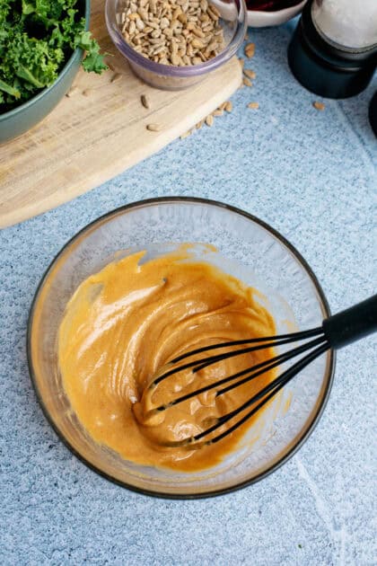 A whisk mixing a smooth and creamy peanut dressing in a glass bowl, with sunflower seeds and fresh ingredients on a wooden board in the background.