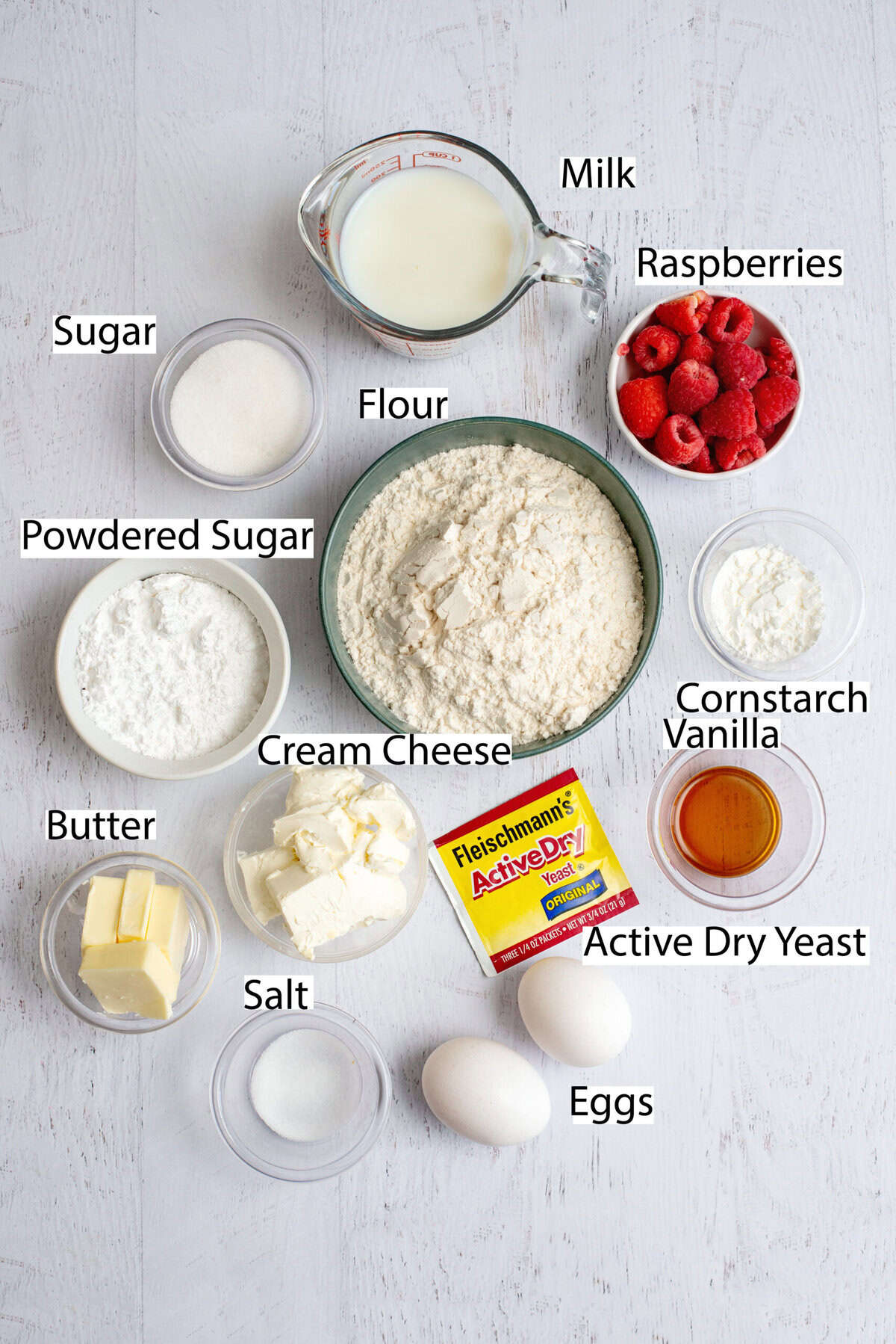 A flat lay of labeled ingredients for raspberry sweet rolls, including flour, sugar, butter, eggs, milk, raspberries, and active dry yeast.