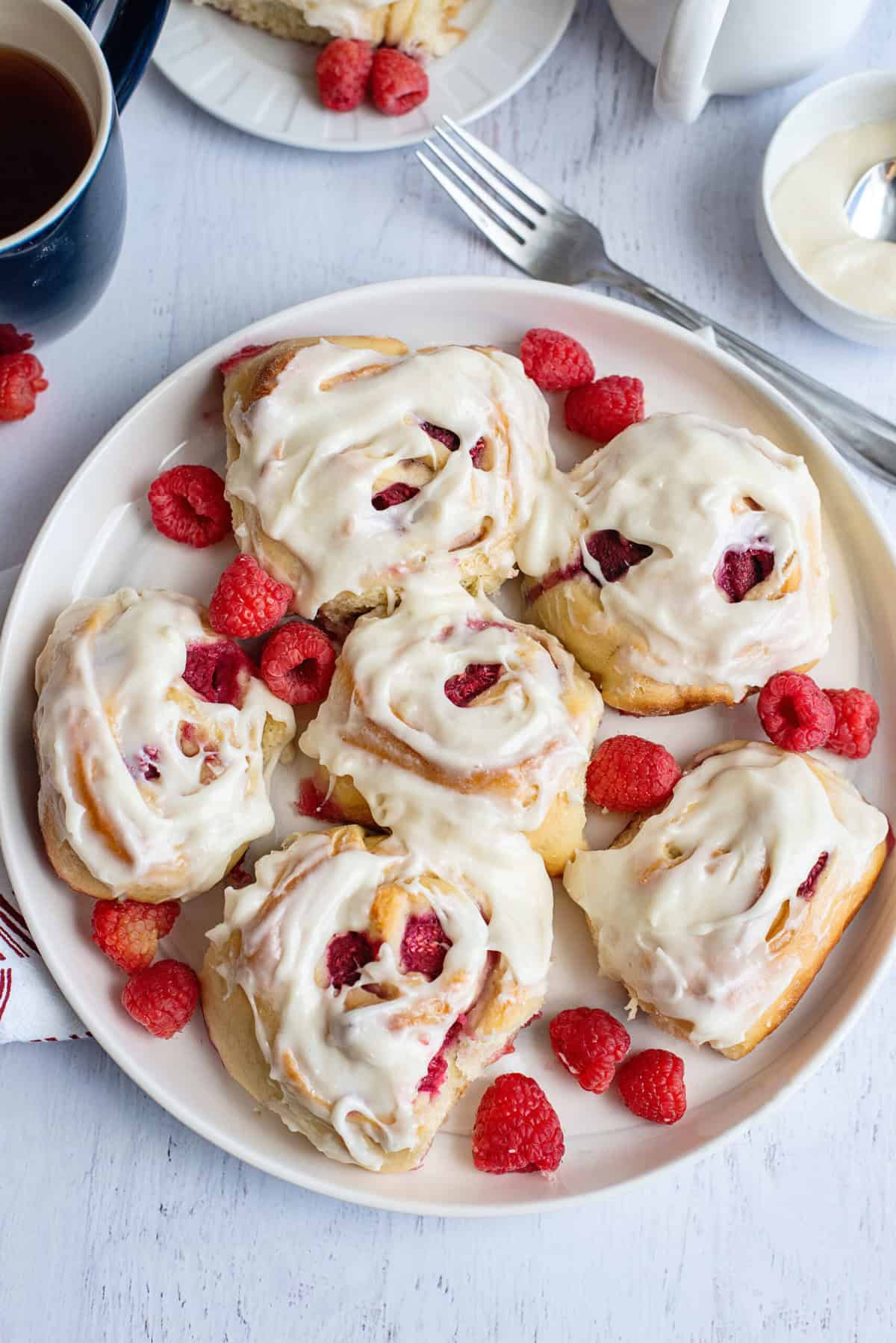 A plate of freshly baked raspberry sweet rolls topped with a thick cream cheese icing sits on a white wooden surface. The rolls are filled with juicy raspberries, and additional fresh raspberries are scattered around the plate. In the background, a fork, a cup of tea, and a small bowl of extra icing accompany the dish, creating a cozy and inviting setting.