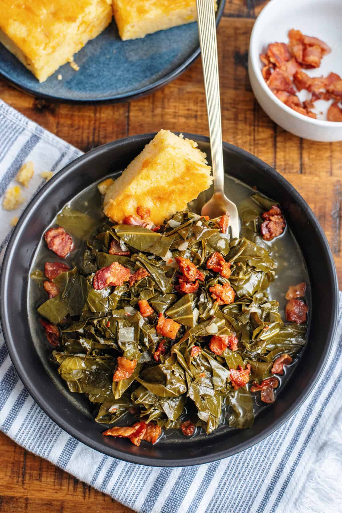 A serving of collard greens in a black bowl with bacon and cornbread.